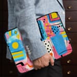 Laptop sleeve with a vibrant abstract design featuring shapes, lines, and patterns in blue, pink, yellow, and black. the person holding the sleeve