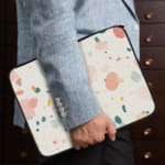 Pastel terrazzo-style laptop sleeve with pink, yellow, blue, and gray shapes on a light background, the person holding the sleeve