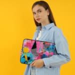 Laptop sleeve with a vibrant abstract design featuring blue, pink, purple, yellow, and black hues, secured with a zip closure. the person holding the sleeve