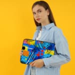 Laptop sleeve with a bright blue background featuring colorful abstract geometric shapes in yellow, pink, orange, black, and red, with zip closure. the person holding the sleeve