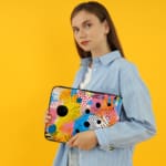 Laptop sleeve with vibrant abstract design in yellow, blue, pink, and black with circles, lines, and dots. the person holding the sleeve