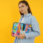 Laptop sleeve with a vibrant abstract design featuring shapes, lines, and patterns in blue, pink, yellow, and black. the person holding the sleeve