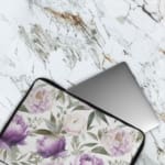 Elegant floral laptop sleeve with purple flowers and green leaves on a soft, light background. The laptop sleeve covers the laptop