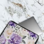 Elegant floral laptop sleeve with purple flowers and green leaves on a soft, light background. The laptop sleeve covers the laptop