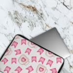 Light laptop sleeve with pink heart, ribbon, and beige mug pattern on a light background. The laptop sleeve covers the laptop