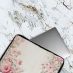 Soft beige laptop sleeve with delicate floral design in pink, green, and purple, featuring a zip closure for secure storage. The laptop sleeve covers the laptop