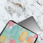 Laptop sleeve with light blue background and pastel popsicle pattern in pink, yellow, blue, and purple, featuring a zip closure. The laptop sleeve covers the laptop