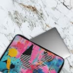 Laptop sleeve with a vibrant abstract design featuring blue, pink, purple, yellow, and black hues, secured with a zip closure. The laptop sleeve covers the laptop