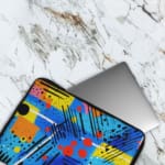 Laptop sleeve with a bright blue background featuring colorful abstract geometric shapes in yellow, pink, orange, black, and red, with zip closure. The laptop sleeve covers the laptop