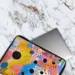 Laptop sleeve with vibrant abstract design in yellow, blue, pink, and black with circles, lines, and dots. The laptop sleeve covers the laptop