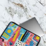 Laptop sleeve with a vibrant abstract design featuring shapes, lines, and patterns in blue, pink, yellow, and black. The laptop sleeve covers the laptop