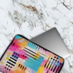 Infidu Bold Abstract Laptop Sleeve featuring bright geometric patterns in pink, blue, yellow, black, and white with a secure zip closure. The laptop sleeve covers the laptop