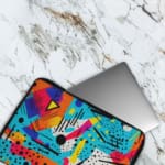 Infidu Vibrant Abstract Laptop Sleeve featuring bold colors like blue, orange, yellow, pink, and purple with geometric shapes and lines. The laptop sleeve covers the laptop