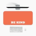 Infidu Be Kind Design Desk Pad featuring bold white text on a bright orange background, perfect for promoting kindness in your workspace. A desk pad under the computer