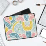 A colorful abstract laptop sleeve featuring pastel pink, blue, yellow, and green curved shapes on a white background with a zip closure. laptop sleeve kept on a plain white background