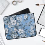 Elegant laptop sleeve with a blue background and white flowers with gray details, featuring small blossoms and leaves, secured with a zip. laptop sleeve kept on a plain white background