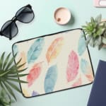 Infidu Pastel Feather Laptop Sleeve with soft fabric in pink, blue, and orange shades, featuring a zip closure for secure laptop protection. the laptop sleeve on the table