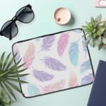 Infidu Pastel Feather Laptop Sleeve with soft fabric in pink, blue, and purple shades, featuring a zip closure for secure laptop protection. the laptop sleeve on the table