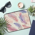 Infidu Pastel Feather Laptop Sleeve with soft pastel feathers in pink, purple, and blue on a light background, shown with office items. the laptop sleeve on the table