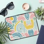 A colorful abstract laptop sleeve featuring pastel pink, blue, yellow, and green curved shapes on a white background with a zip closure. laptop sleeve kept on a plain white background the laptop sleeve on the table