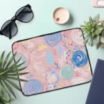 An abstract pastel laptop sleeve in pink, blue, and light green with circular and swirl patterns on a white background with a zip closure. the laptop sleeve on the table