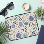 Organic abstract laptop sleeve with hand-drawn patterns in coral, purple, mint green, and black on a white background with a zip closure. the laptop sleeve on the table