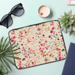 Laptop sleeve with a colorful terrazzo pattern in pink, orange, yellow, green, and black shapes on a white background, with a zip closure. the laptop sleeve on the table