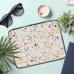 Laptop sleeve with a colorful speckled design in black, orange, blue, and yellow on a light background, featuring a secure zip closure. the laptop sleeve on the table