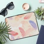 Laptop sleeve with a soft pastel feather design in pink, blue, purple, and orange on a light background, featuring a secure zip closure. the laptop sleeve on the table