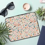 Terrazzo design laptop sleeve with blue, brown, and beige shapes on a light background, placed on a white desk with office supplies. the laptop sleeve on the table