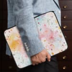 Colorful splatter paint design laptop sleeve with blue, orange, and pink splashes on a light background. the person holding the sleeve