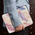 Infidu Pastel Feather Laptop Sleeve with soft pastel feathers in pink, purple, and blue on a light background, shown with office items. the person holding the sleeve