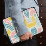 A colorful abstract laptop sleeve featuring pastel pink, blue, yellow, and green curved shapes on a white background with a zip closure. laptop sleeve kept on a plain white background the person holding the sleeve