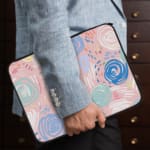 An abstract pastel laptop sleeve in pink, blue, and light green with circular and swirl patterns on a white background with a zip closure. the person holding the sleeve