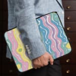 Retro-inspired laptop sleeve with wavy pastel stripes in pink, purple, yellow, green, and blue, covering the surface with a zip closure. the person holding the sleeve