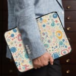 Laptop sleeve with a pastel bubble pattern in light blue, coral pink, and peach circles on a white background, secured with a zip closure. the person holding the sleeve