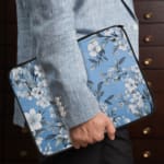 Elegant laptop sleeve with a blue background and white flowers with gray details, featuring small blossoms and leaves, secured with a zip. the person holding the sleeve