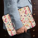 Laptop sleeve with a colorful terrazzo pattern in pink, orange, yellow, green, and black shapes on a white background, with a zip closure. the person holding the sleeve