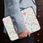 Laptop sleeve with colorful polka dots in pastel shades of pink, blue, yellow, and purple on a light background, with a secure zip closure. the person holding the sleeve