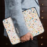 Laptop sleeve with a colorful speckled design in black, orange, blue, and yellow on a light background, featuring a secure zip closure. the person holding the sleeve