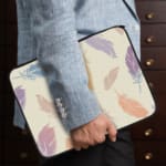 Laptop sleeve with a soft pastel feather design in pink, blue, purple, and orange on a light background, featuring a secure zip closure. the person holding the sleeve