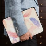 Laptop sleeve with a soft pastel feather design in pink, blue, purple, and orange on a light background, featuring a secure zip closure. the person holding the sleeve