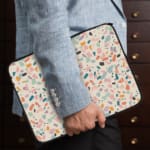 Terrazzo design laptop sleeve with blue, brown, and beige shapes on a light background, placed on a white desk with office supplies. the person holding the sleeve