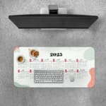 Infidu Calendar Design Desk Pad displaying a 2025 calendar layout with a light background and abstract red and gray accents. Desk Pad with Keyboard and Mouse on top of the desk pad