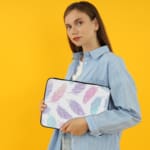 Infidu Pastel Feather Laptop Sleeve with soft fabric in pink, blue, and purple shades, featuring a zip closure for secure laptop protection. the person holding the sleeve