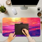 Infidu Calming Sunset Reflection Design Desk Pad featuring a sunset over water in soft shades of pink, purple, orange, and yellow. A writing pad on top of the desk pad