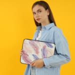 Infidu Pastel Feather Laptop Sleeve with soft pastel feathers in pink, purple, and blue on a light background, shown with office items. the person holding the sleeve