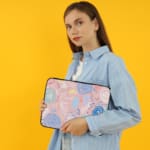 An abstract pastel laptop sleeve in pink, blue, and light green with circular and swirl patterns on a white background with a zip closure. the person holding the sleeve