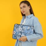 Elegant laptop sleeve with a blue background and white flowers with gray details, featuring small blossoms and leaves, secured with a zip. the person holding the sleeve