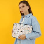 Laptop sleeve with a colorful speckled design in black, orange, blue, and yellow on a light background, featuring a secure zip closure. the person holding the sleeve